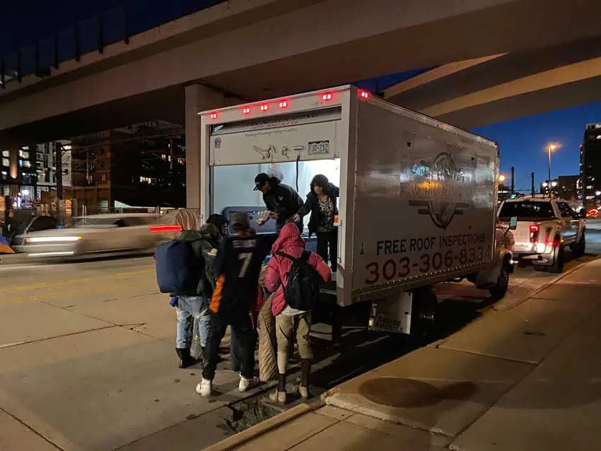 Neighbors lining up for food at the ARCM Roofing truck under a Denver overpass at night